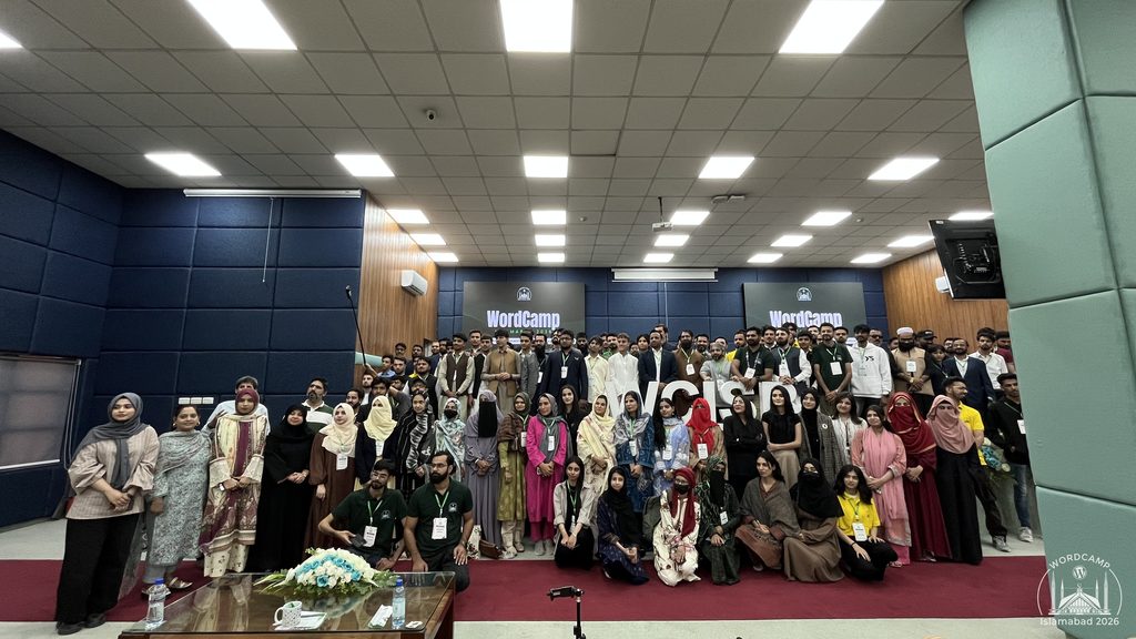 A large group of diverse individuals posing together on stage at WordCamp Islamabad 2026, with a backdrop featuring the WordCamp logo and seating in front.